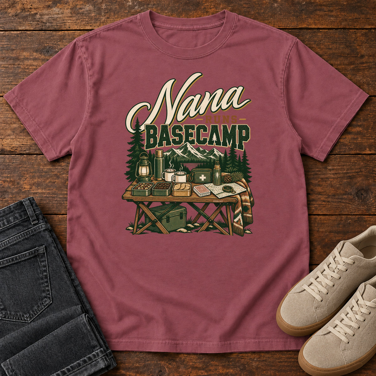 Brick Nana Runs Basecamp T-Shirt with a camp table with mugs, lantern, snacks and supplies beneath mountains; front flat lay on wood.