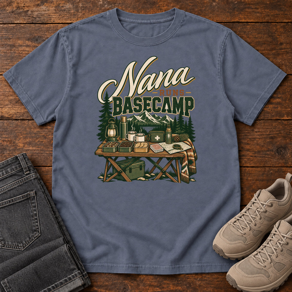 Denim Nana Runs Basecamp T-Shirt with a camp table with mugs, lantern, snacks and supplies beneath mountains; front flat lay on wood.