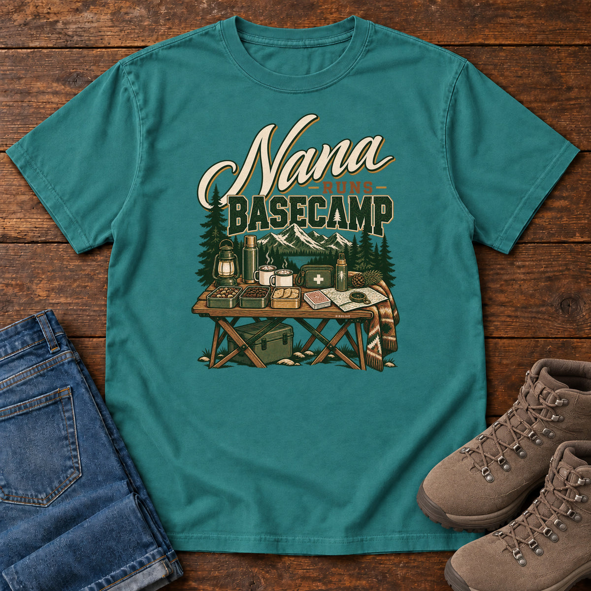 Emerald Nana Runs Basecamp T-Shirt with a camp table with mugs, lantern, snacks and supplies beneath mountains; front flat lay on wood.