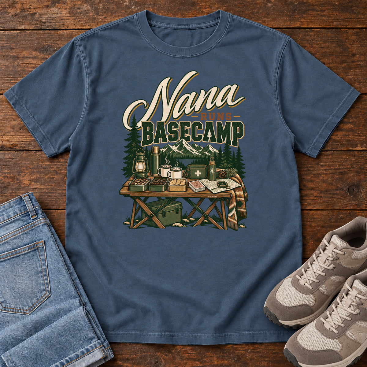 Midnight Nana Runs Basecamp T-Shirt with a camp table with mugs, lantern, snacks and supplies beneath mountains; front flat lay on wood.