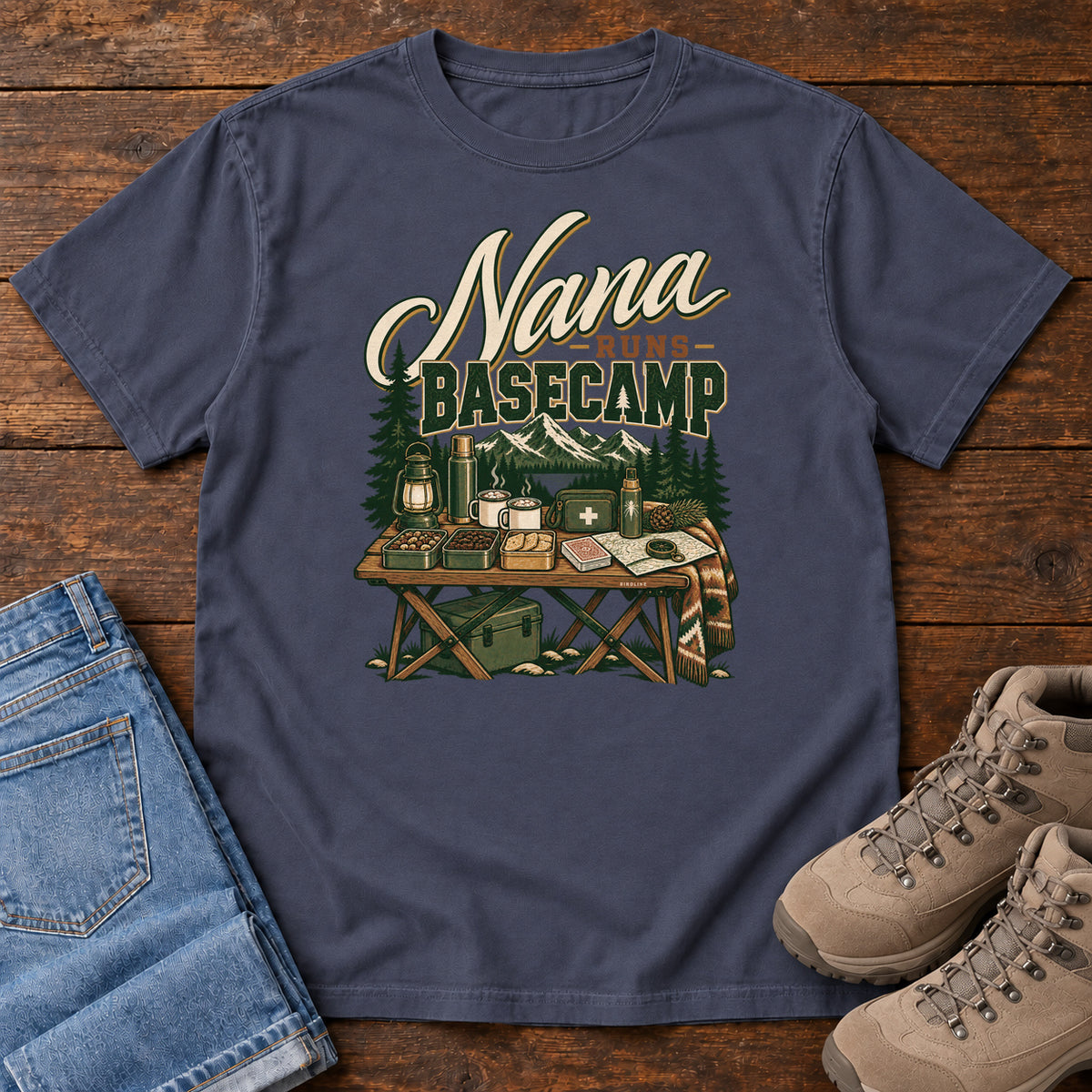 Navy Nana Runs Basecamp T-Shirt with a camp table with mugs, lantern, snacks and supplies beneath mountains; front flat lay on wood.