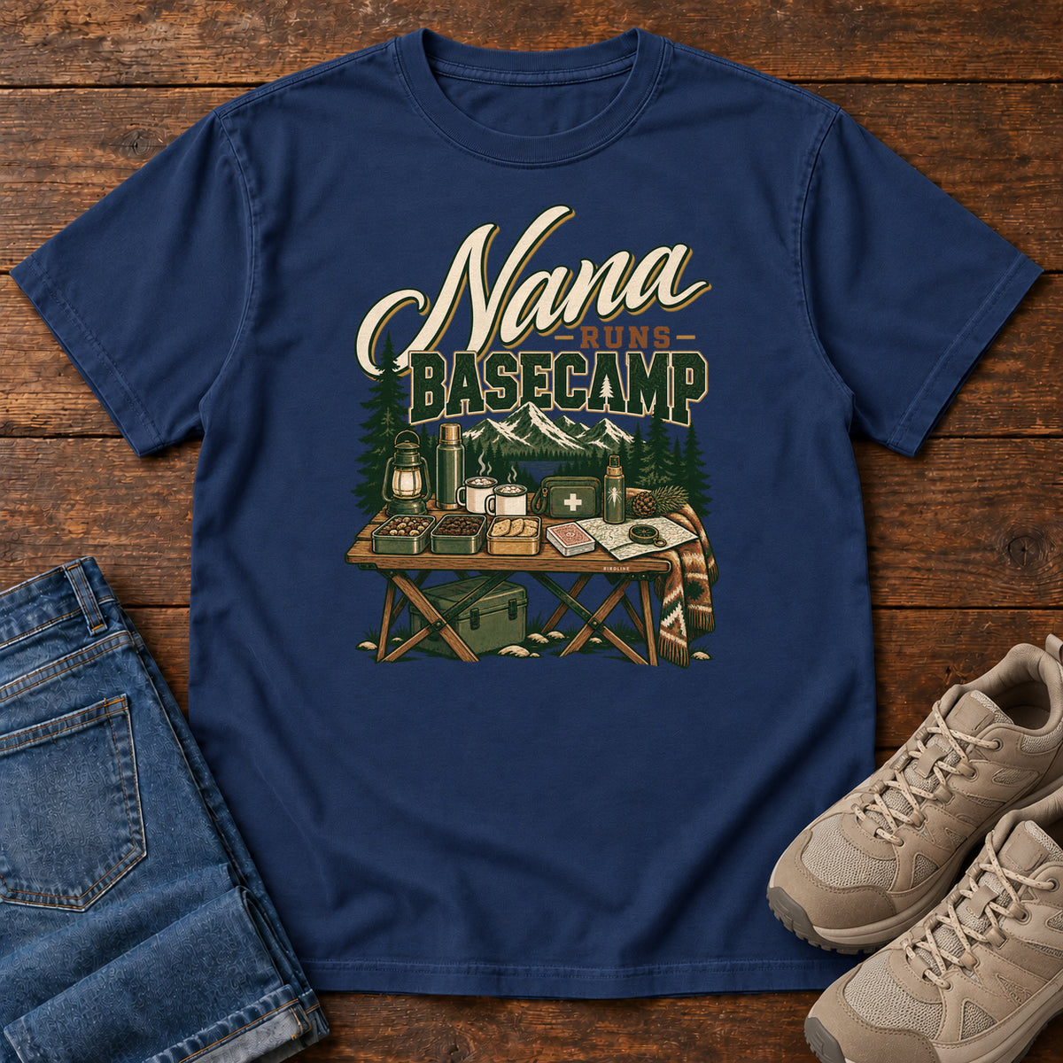 True Navy Nana Runs Basecamp T-Shirt with a camp table with mugs, lantern, snacks and supplies beneath mountains; front flat lay on wood.