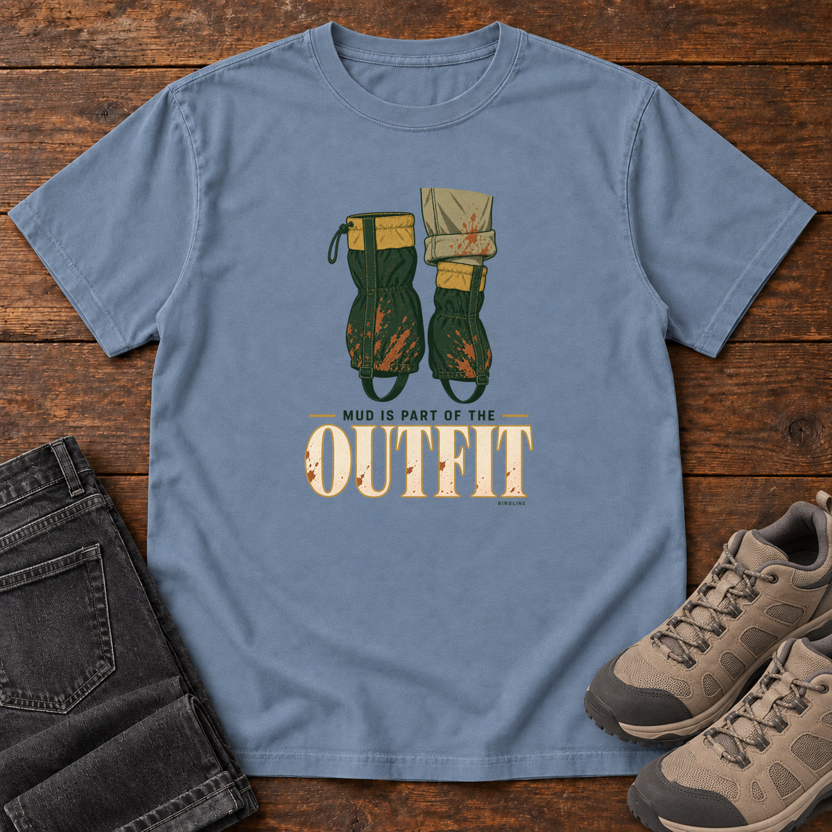 Blue Jean Mud Is Part of the Outfit T-Shirt with mud-splashed green boots and cream lettering, front flat lay on wood.