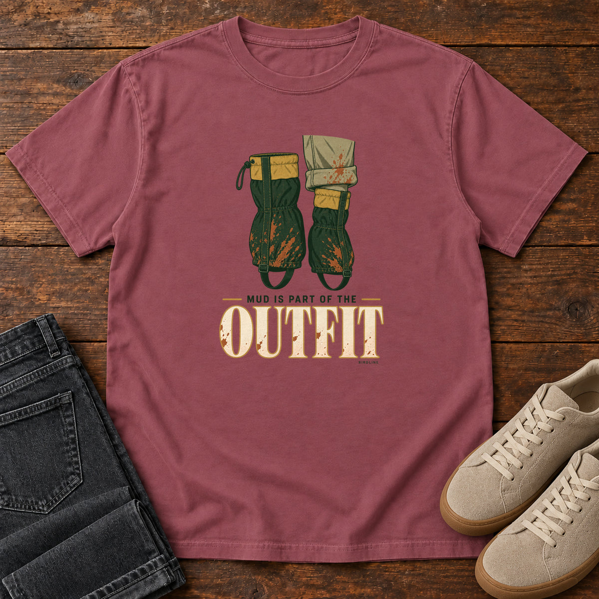 Brick Mud Is Part of the Outfit T-Shirt with mud-splashed green boots and cream lettering, front flat lay on wood.