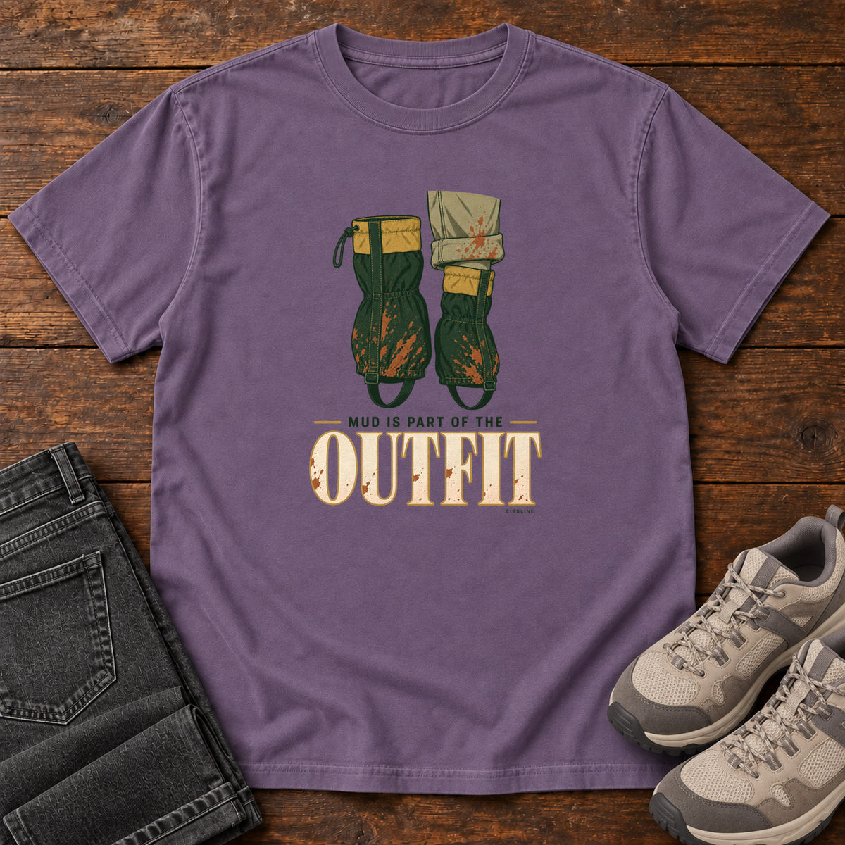 Grape Mud Is Part of the Outfit T-Shirt with mud-splashed green boots and cream lettering, front flat lay on wood.