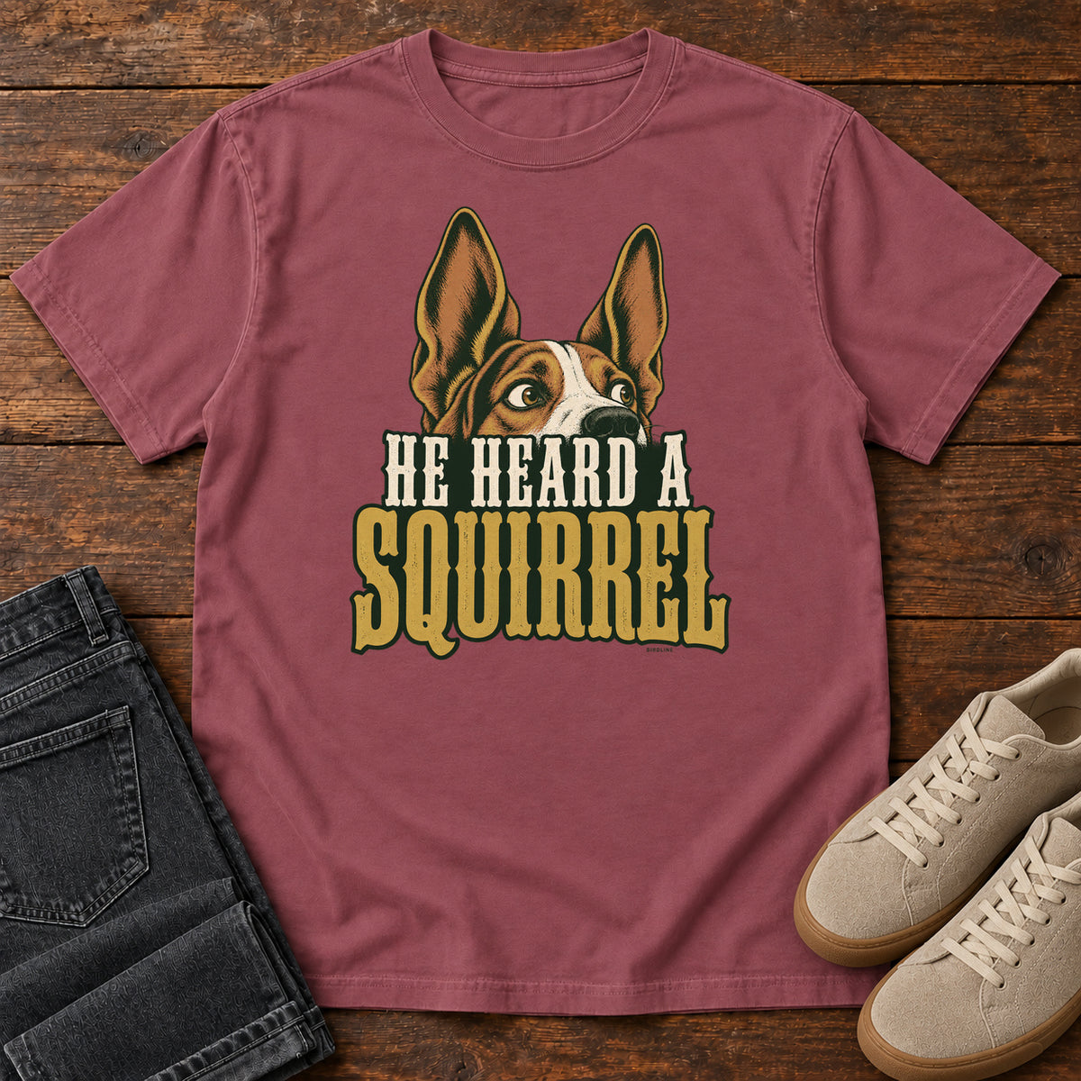 Brick He Heard a Squirrel T-Shirt with an alert dog with upright ears above He Heard a Squirrel lettering; front flat lay on wood.