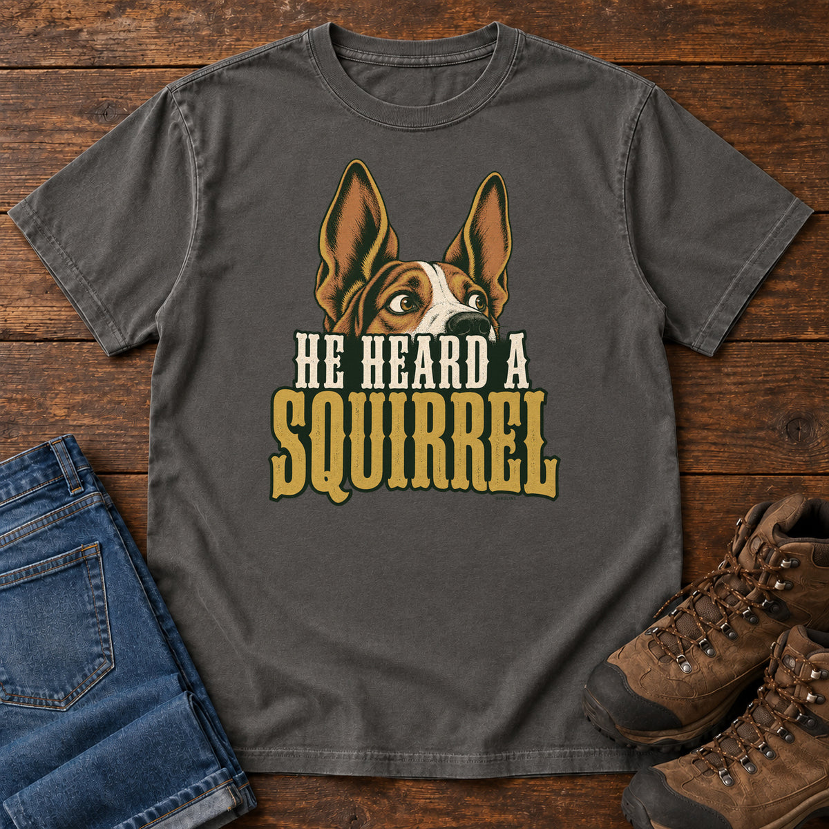 Pepper He Heard a Squirrel T-Shirt with an alert dog with upright ears above He Heard a Squirrel lettering; front flat lay on wood.
