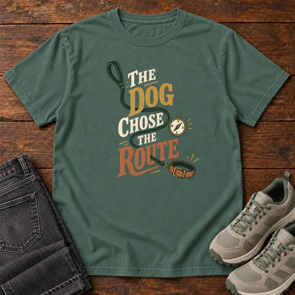 Blue Spruce The Dog Chose the Route T-Shirt with a green leash, dog collar and compass around stacked lettering; front flat lay on wood.