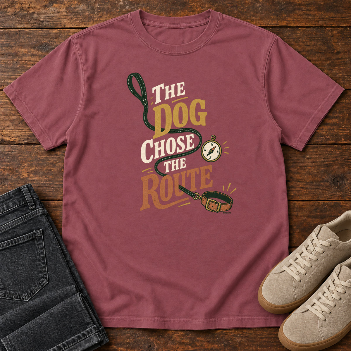Brick The Dog Chose the Route T-Shirt with a green leash, dog collar and compass around stacked lettering; front flat lay on wood.