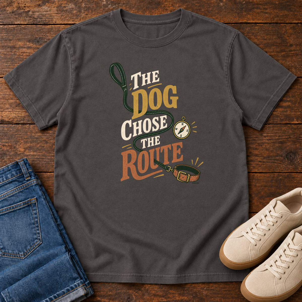 Graphite The Dog Chose the Route T-Shirt with a green leash, dog collar and compass around stacked lettering; front flat lay on wood.