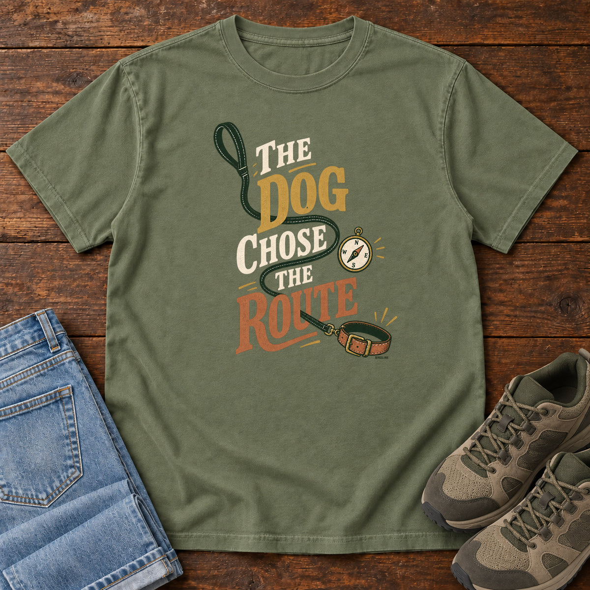 Moss The Dog Chose the Route T-Shirt with a green leash, dog collar and compass around stacked lettering; front flat lay on wood.