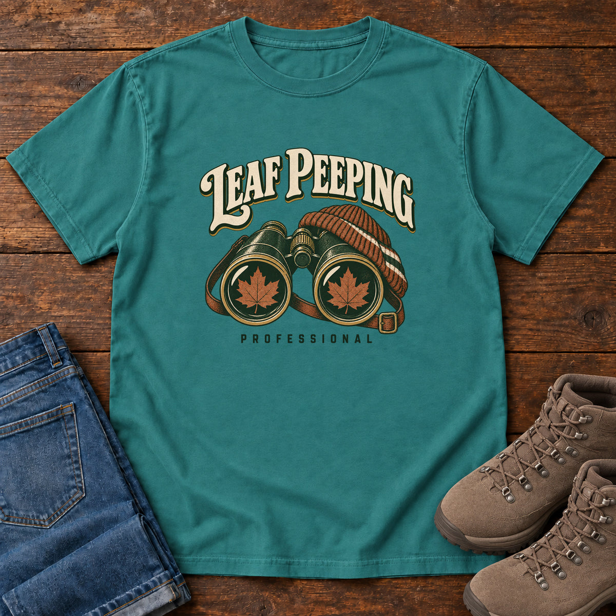 Leaf Peeping Professional T-Shirt in Emerald, front laid flat on wood, featuring binoculars with orange leaves in the lenses and a striped knit hat beneath cream lettering.