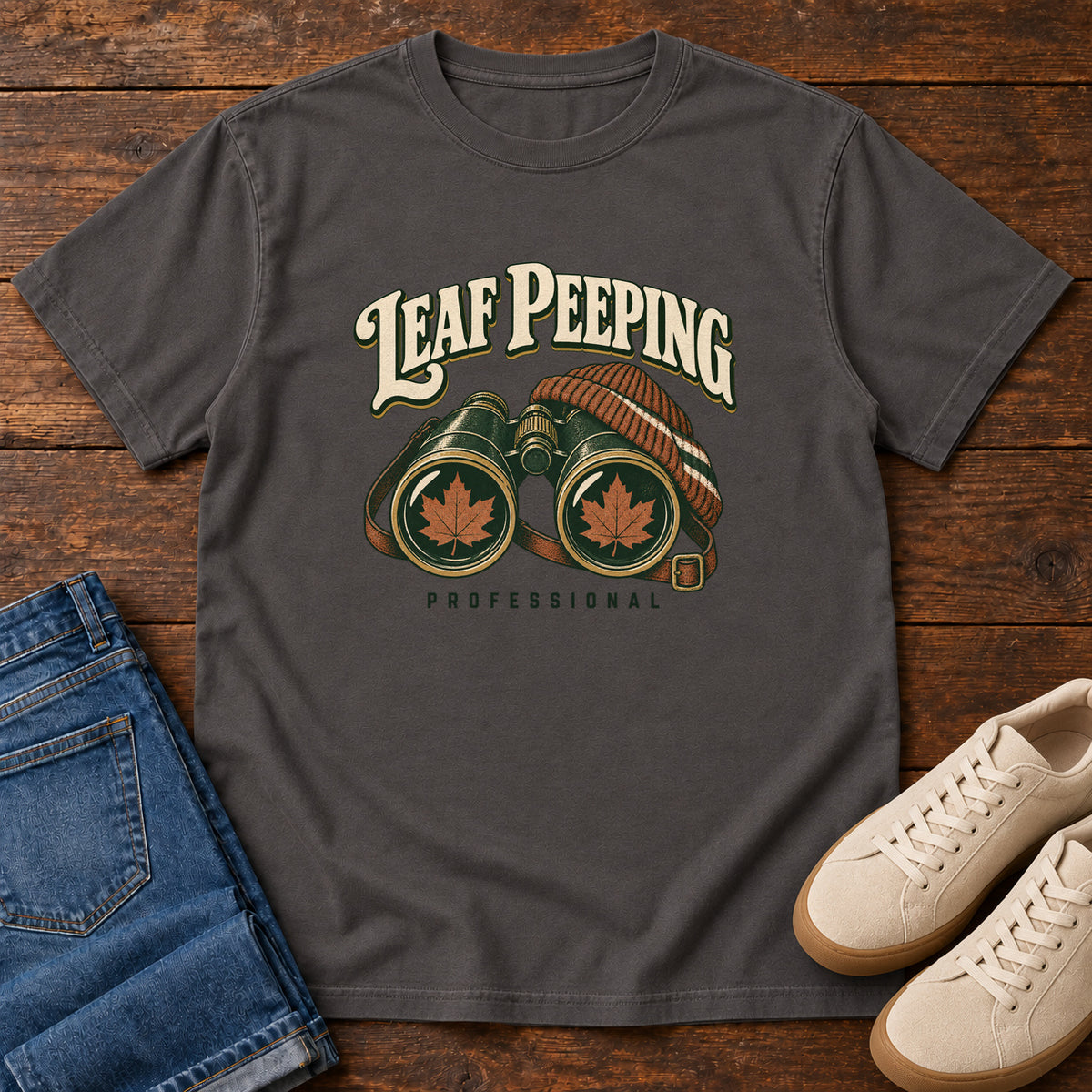 Leaf Peeping Professional T-Shirt in Graphite, front laid flat on wood, featuring binoculars with orange leaves in the lenses and a striped knit hat beneath cream lettering.