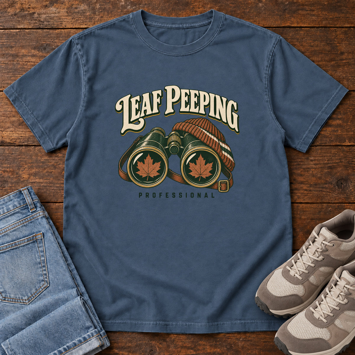 Leaf Peeping Professional T-Shirt in Midnight, front laid flat on wood, featuring binoculars with orange leaves in the lenses and a striped knit hat beneath cream lettering.