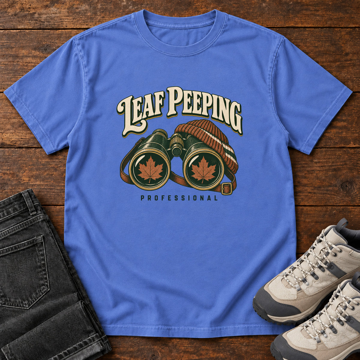 Leaf Peeping Professional T-Shirt in Mystic Blue, front laid flat on wood, featuring binoculars with orange leaves in the lenses and a striped knit hat beneath cream lettering.