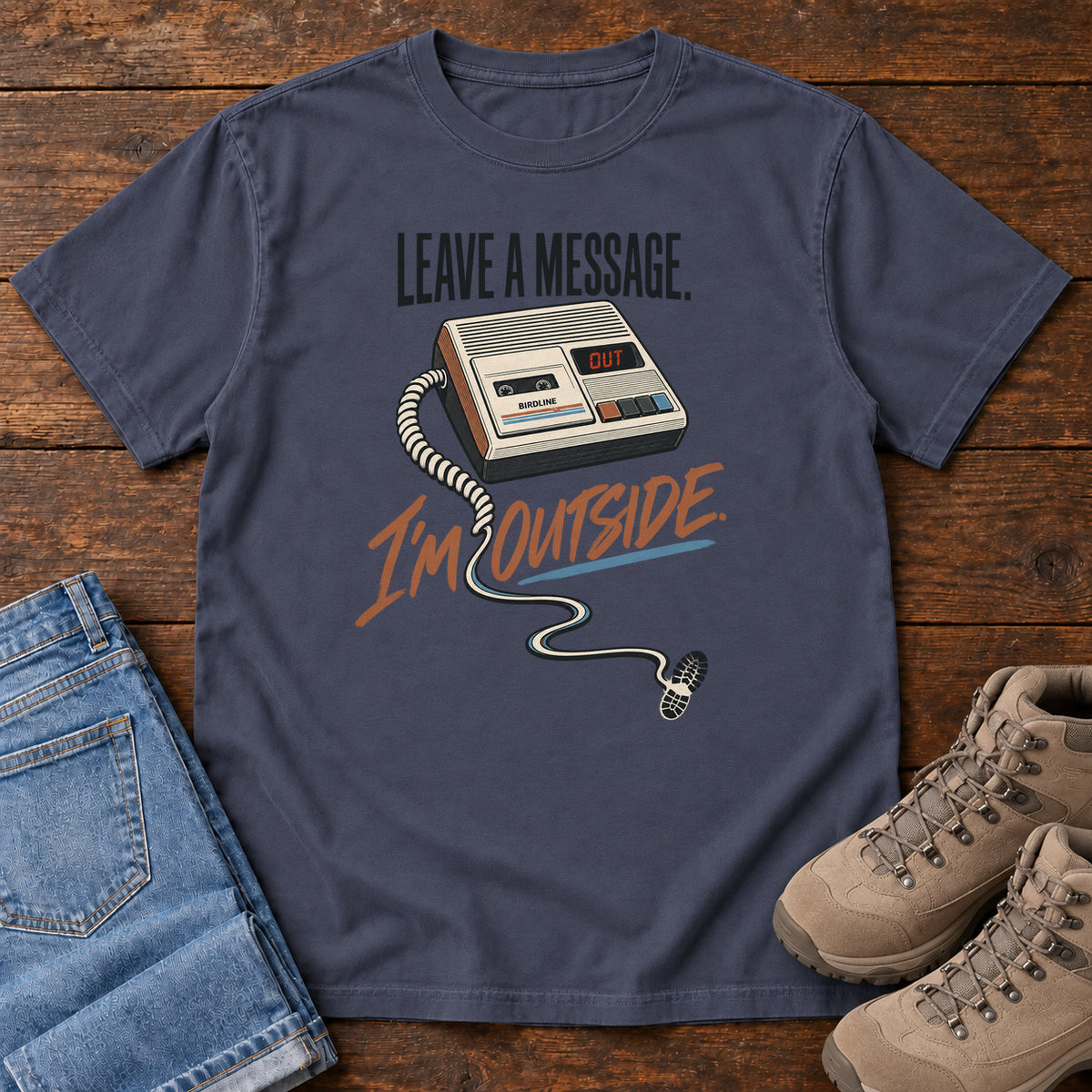 Navy Leave a Message I'm Outside T-Shirt with a retro answering machine, trailing cord and orange script, front flat lay on wood.