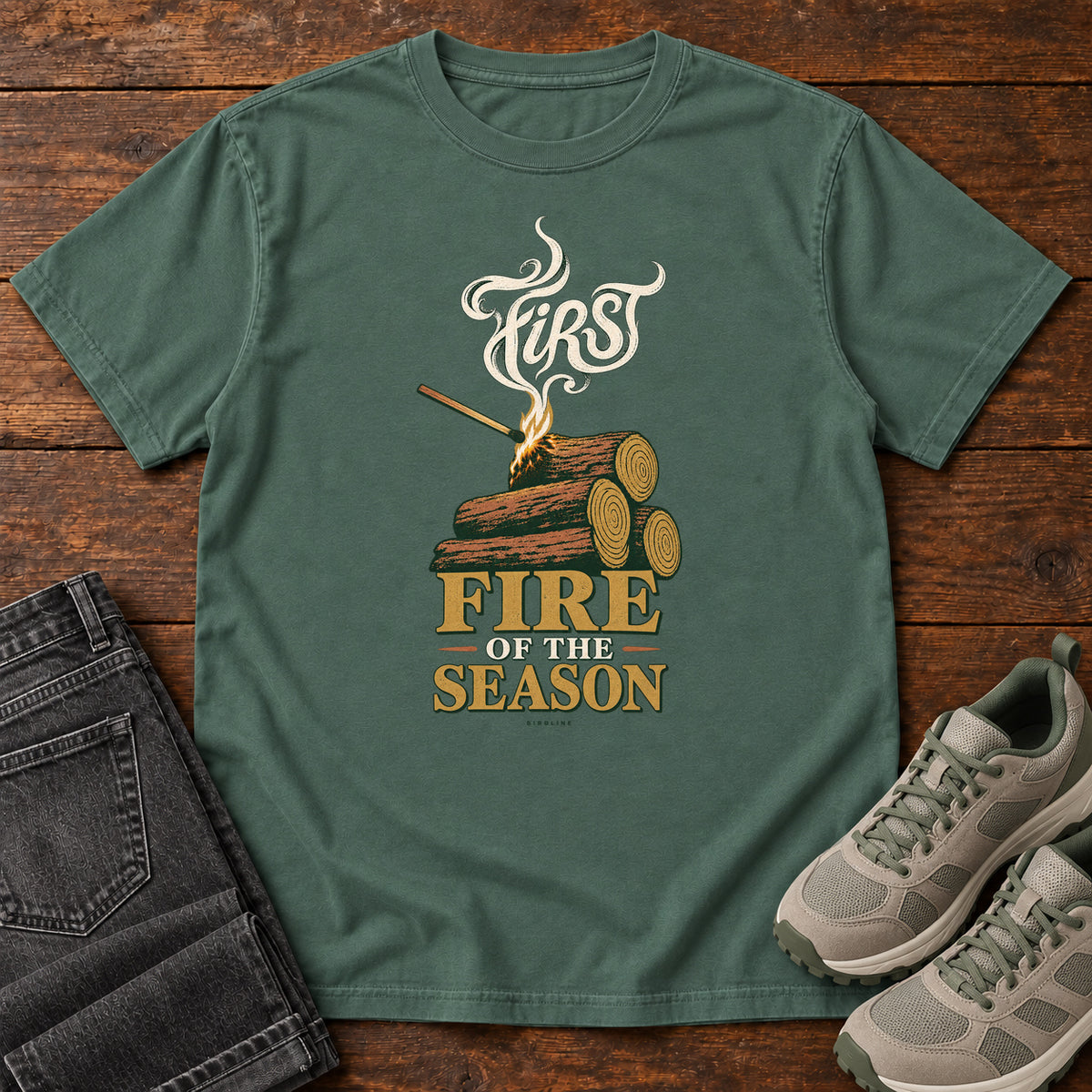 Blue Spruce First Fire of the Season T-Shirt with stacked logs, a small flame and curling smoke around First Fire of the Season lettering; front flat lay on wood.