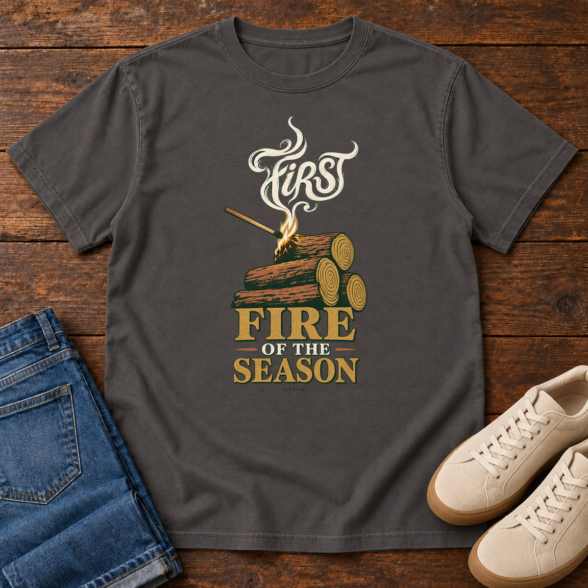 Graphite First Fire of the Season T-Shirt with stacked logs, a small flame and curling smoke around First Fire of the Season lettering; front flat lay on wood.