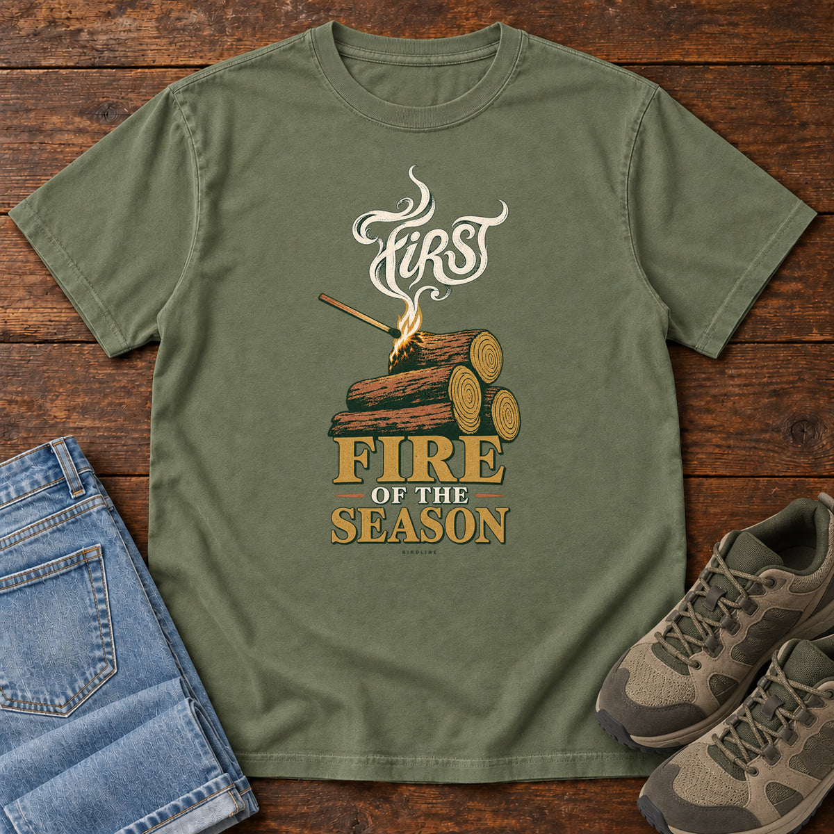 Moss First Fire of the Season T-Shirt with stacked logs, a small flame and curling smoke around First Fire of the Season lettering; front flat lay on wood.