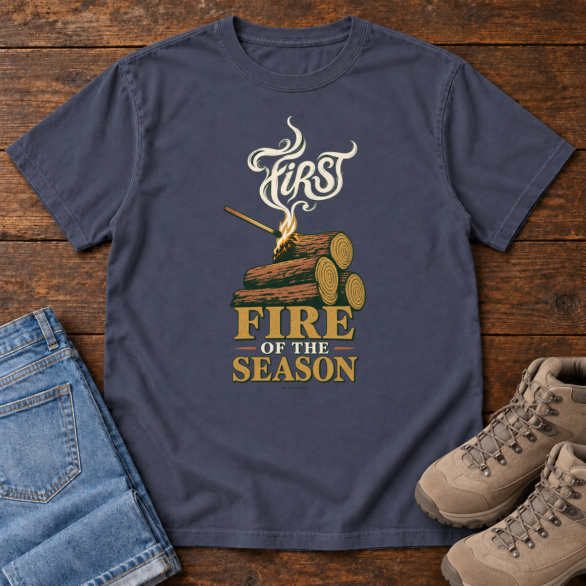 Navy First Fire of the Season T-Shirt with stacked logs, a small flame and curling smoke around First Fire of the Season lettering; front flat lay on wood.