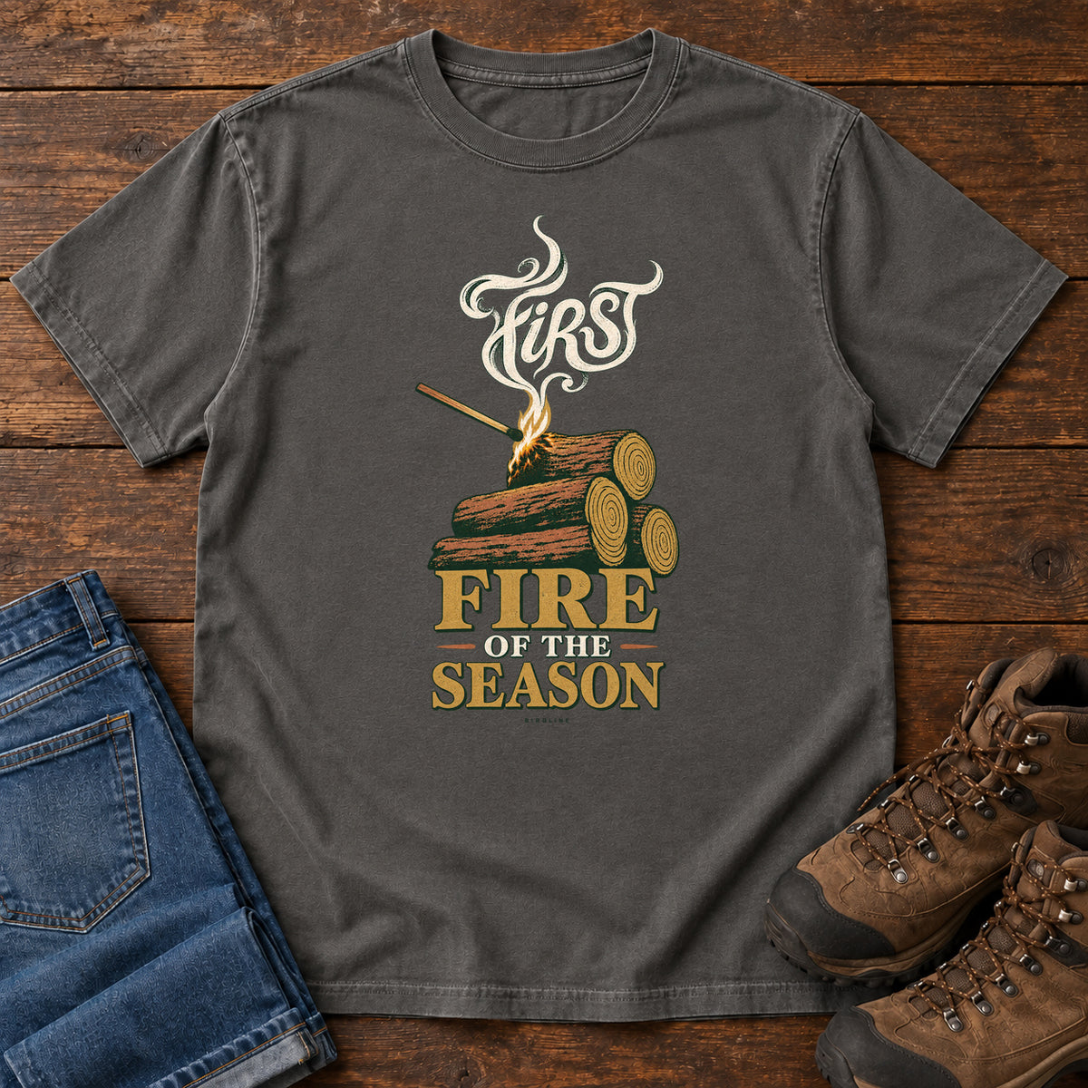 Pepper First Fire of the Season T-Shirt with stacked logs, a small flame and curling smoke around First Fire of the Season lettering; front flat lay on wood.