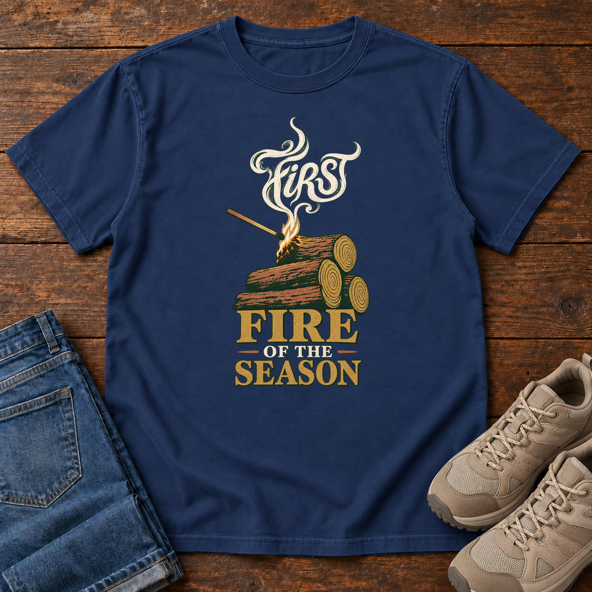 True Navy First Fire of the Season T-Shirt with stacked logs, a small flame and curling smoke around First Fire of the Season lettering; front flat lay on wood.