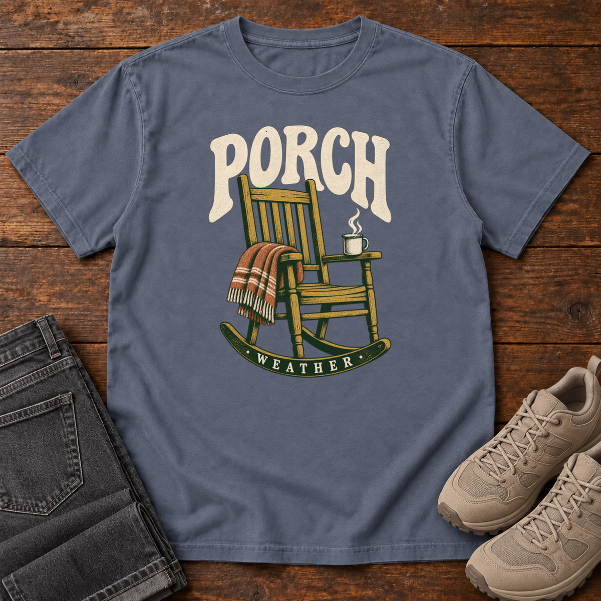 Denim Porch Weather T-Shirt with a wooden rocking chair with a striped blanket and steaming mug; front flat lay on wood.