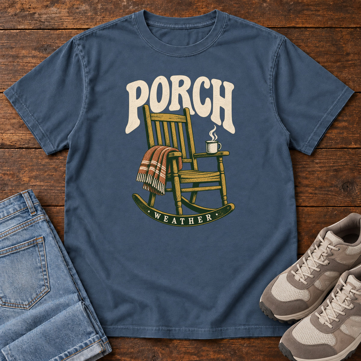 Midnight Porch Weather T-Shirt with a wooden rocking chair with a striped blanket and steaming mug; front flat lay on wood.