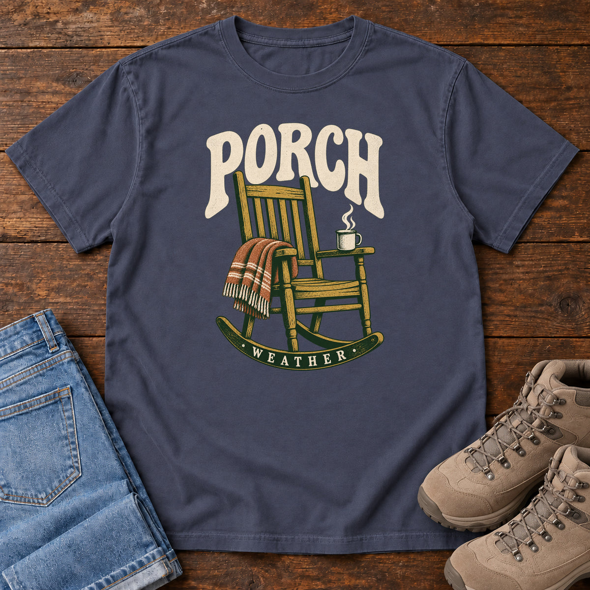Navy Porch Weather T-Shirt with a wooden rocking chair with a striped blanket and steaming mug; front flat lay on wood.