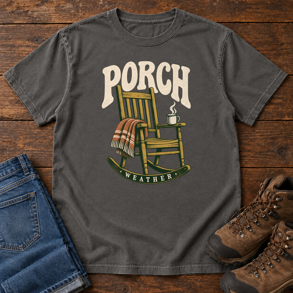 Pepper Porch Weather T-Shirt with a wooden rocking chair with a striped blanket and steaming mug; front flat lay on wood.