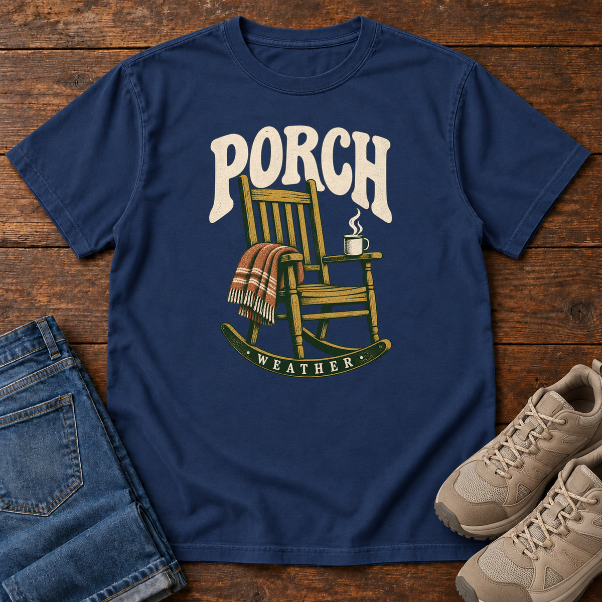 True Navy Porch Weather T-Shirt with a wooden rocking chair with a striped blanket and steaming mug; front flat lay on wood.