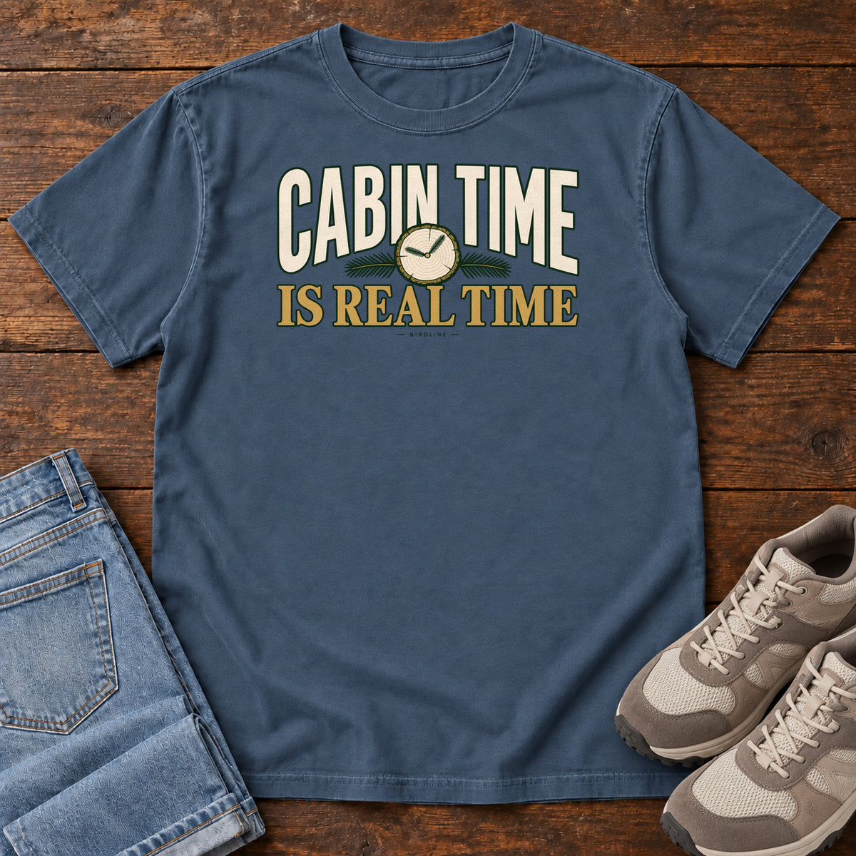 Midnight Cabin Time Is Real Time T-Shirt with cream and gold lettering surrounding a small clock illustration, front flat lay on wood.