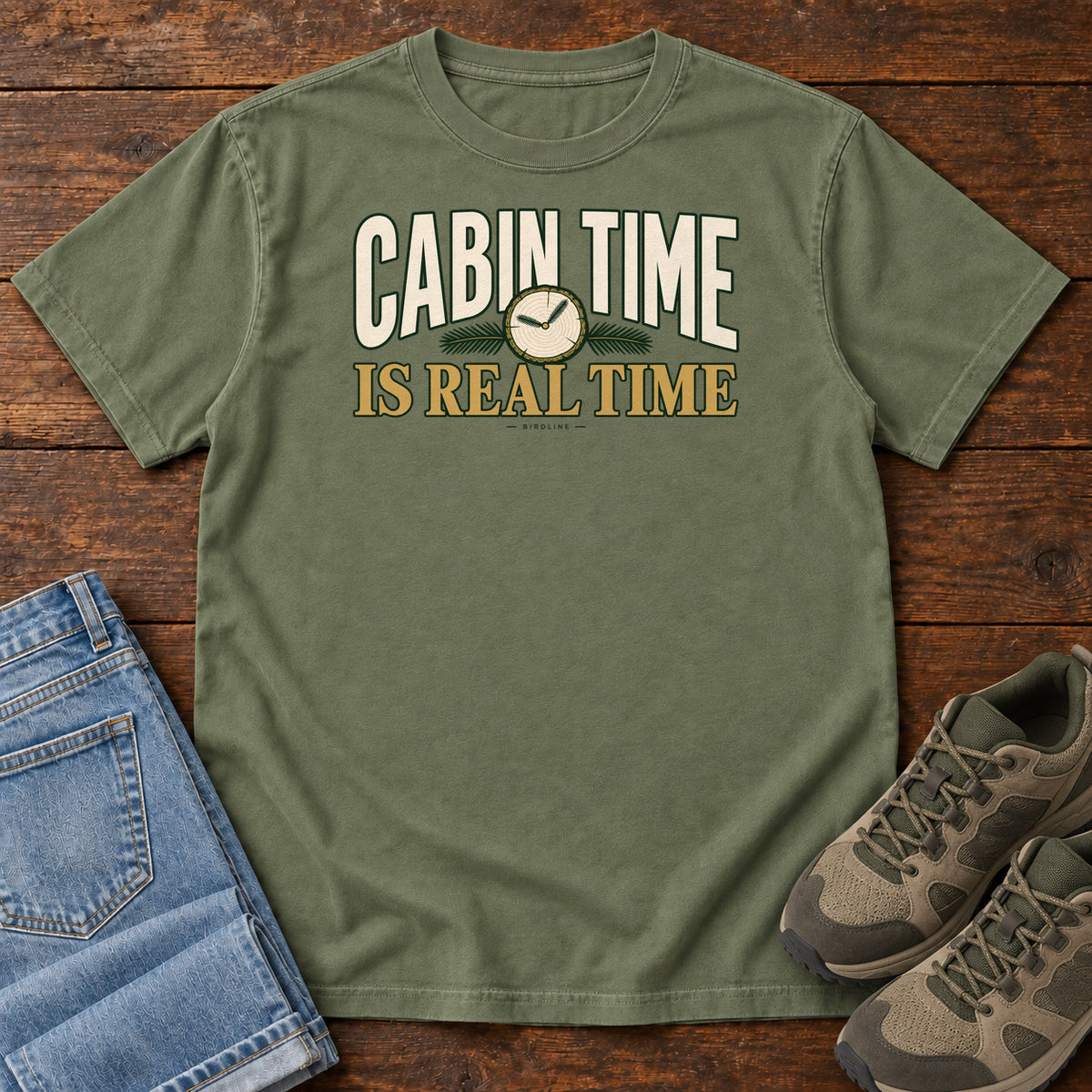 Moss Cabin Time Is Real Time T-Shirt with cream and gold lettering surrounding a small clock illustration, front flat lay on wood.