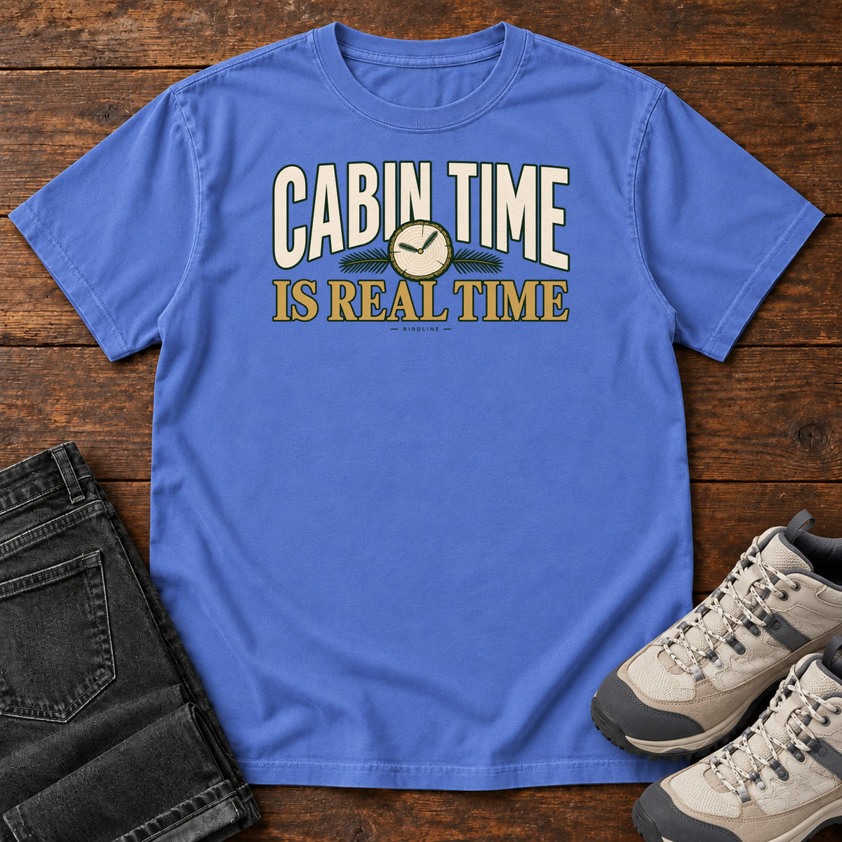 Mystic Blue Cabin Time Is Real Time T-Shirt with cream and gold lettering surrounding a small clock illustration, front flat lay on wood.