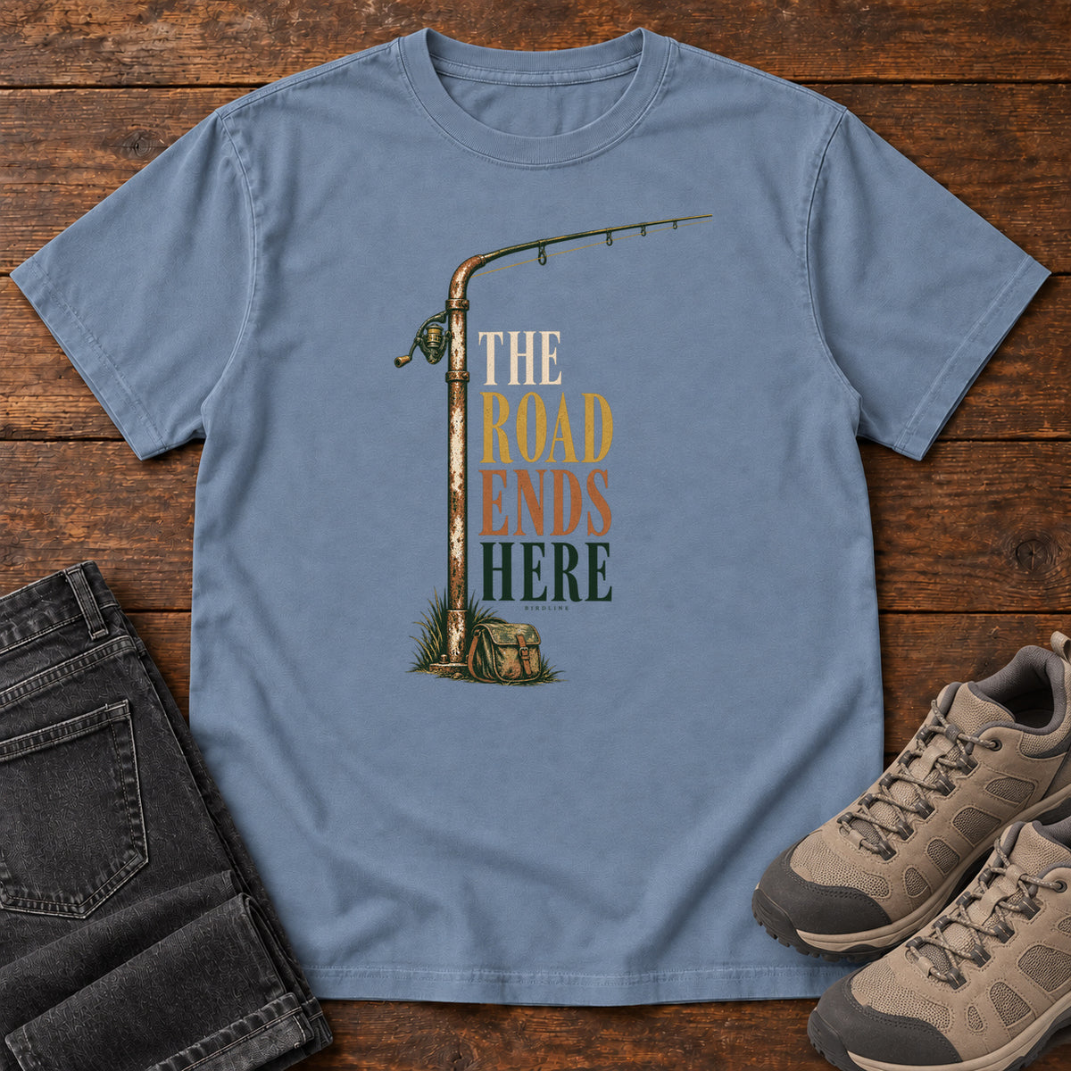 The Road Ends Here T-Shirt in Blue Jean, front laid flat on wood, featuring a fishing rod beside stacked earth-tone slogan lettering.
