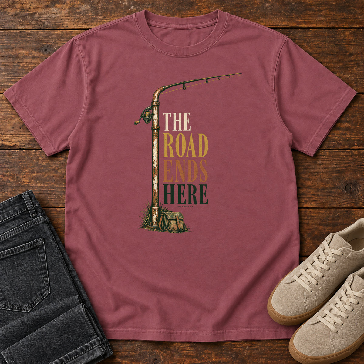 The Road Ends Here T-Shirt in Brick, front laid flat on wood, featuring a fishing rod beside stacked earth-tone slogan lettering.