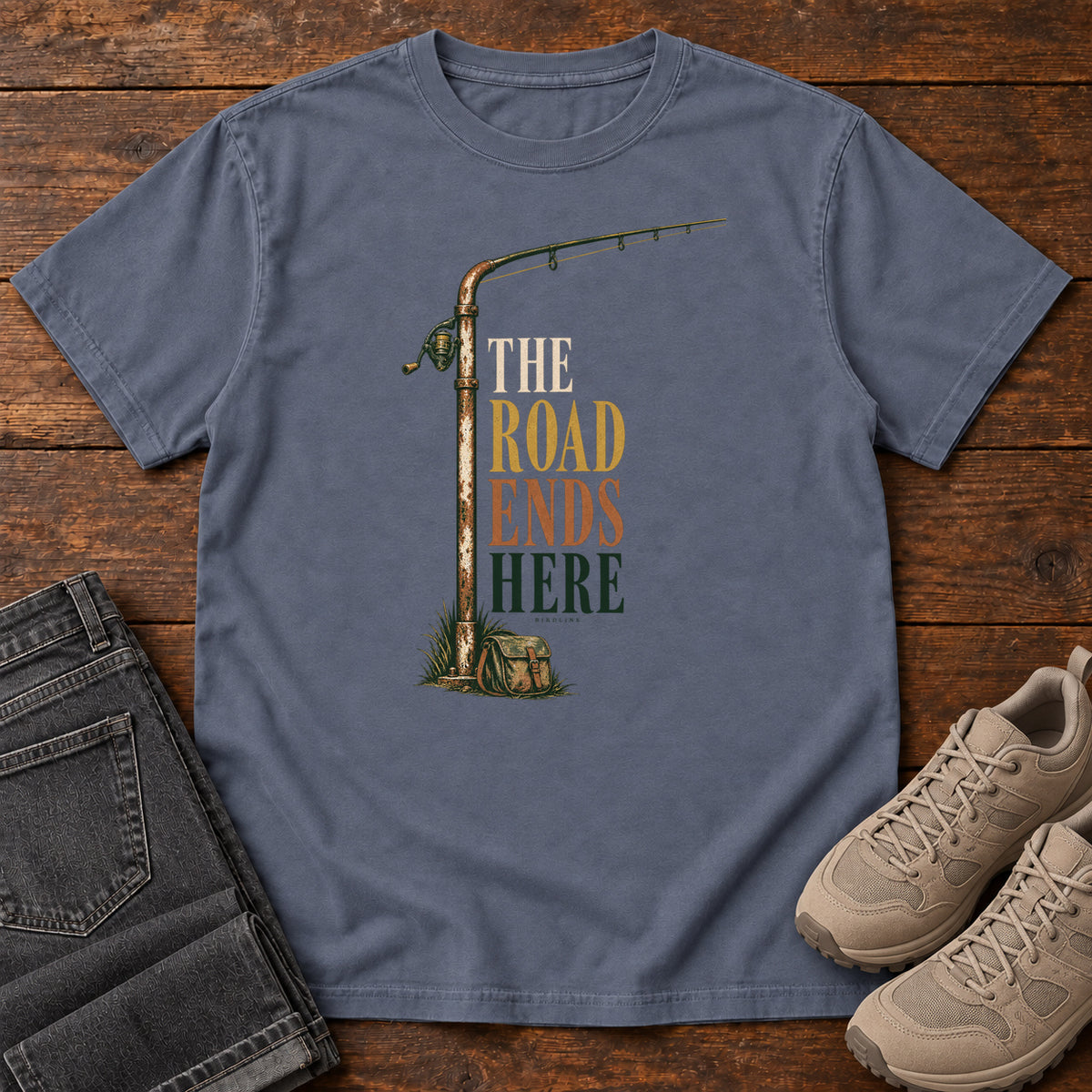 The Road Ends Here T-Shirt in Denim, front laid flat on wood, featuring a fishing rod beside stacked earth-tone slogan lettering.