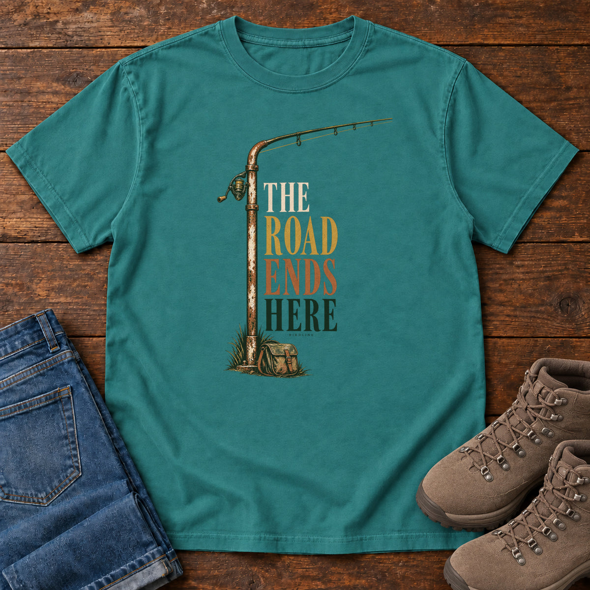 The Road Ends Here T-Shirt in Emerald, front laid flat on wood, featuring a fishing rod beside stacked earth-tone slogan lettering.
