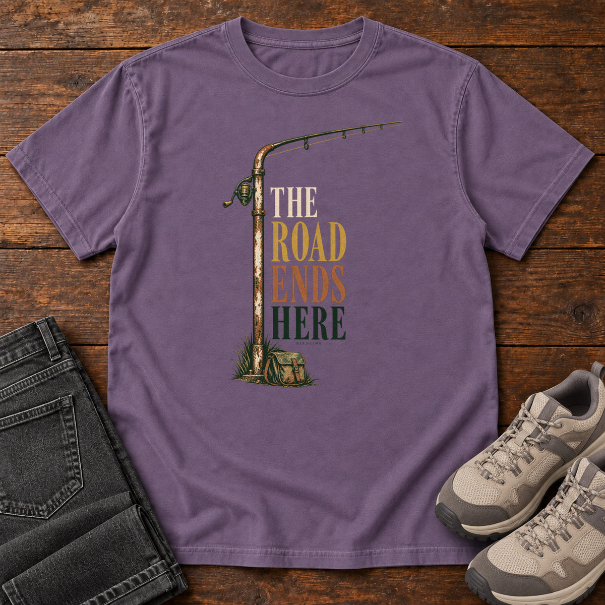 The Road Ends Here T-Shirt in Grape, front laid flat on wood, featuring a fishing rod beside stacked earth-tone slogan lettering.