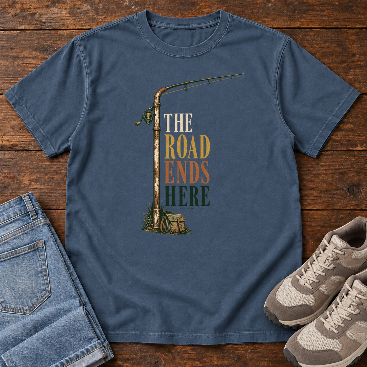 The Road Ends Here T-Shirt in Midnight, front laid flat on wood, featuring a fishing rod beside stacked earth-tone slogan lettering.