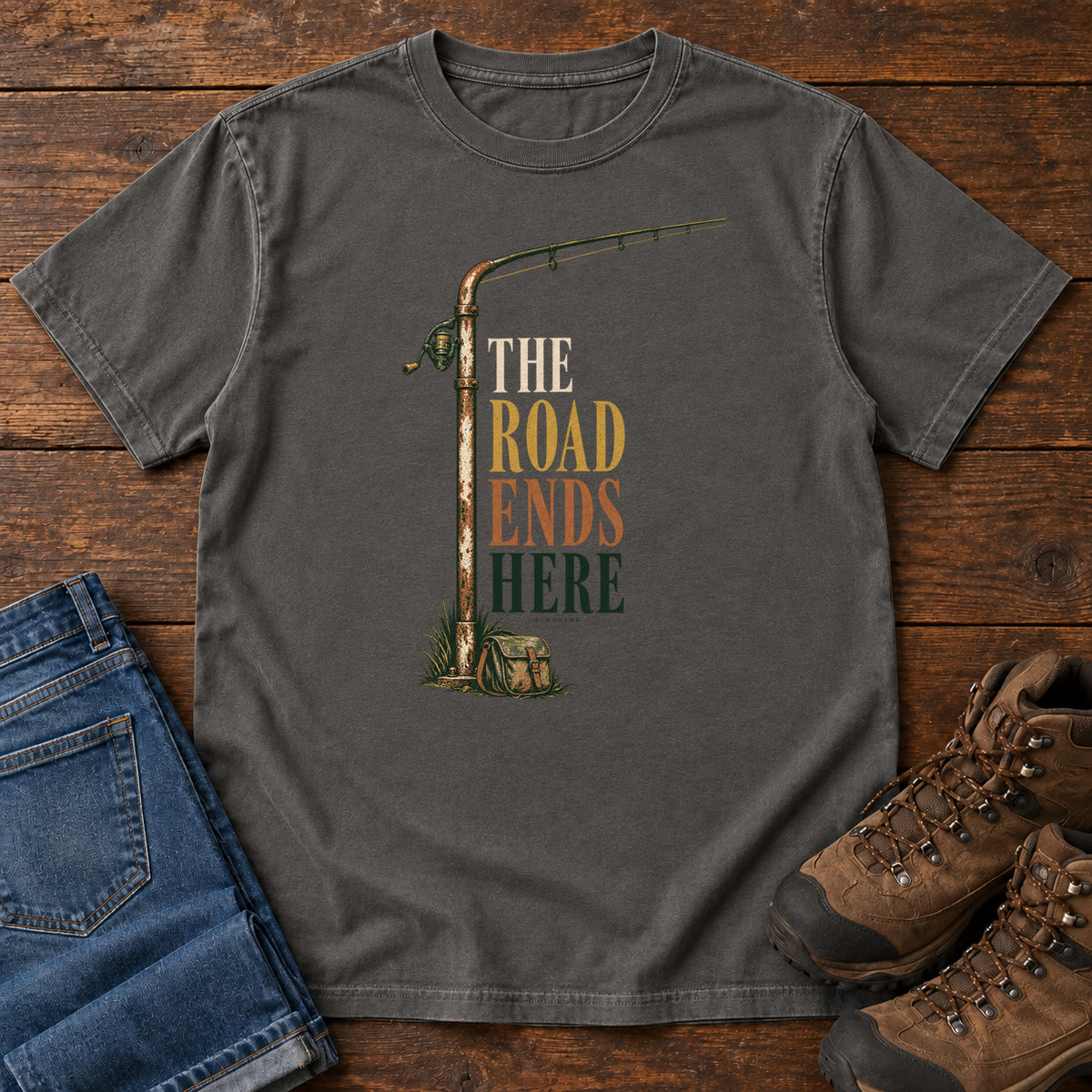 The Road Ends Here T-Shirt in Pepper, front laid flat on wood, featuring a fishing rod beside stacked earth-tone slogan lettering.