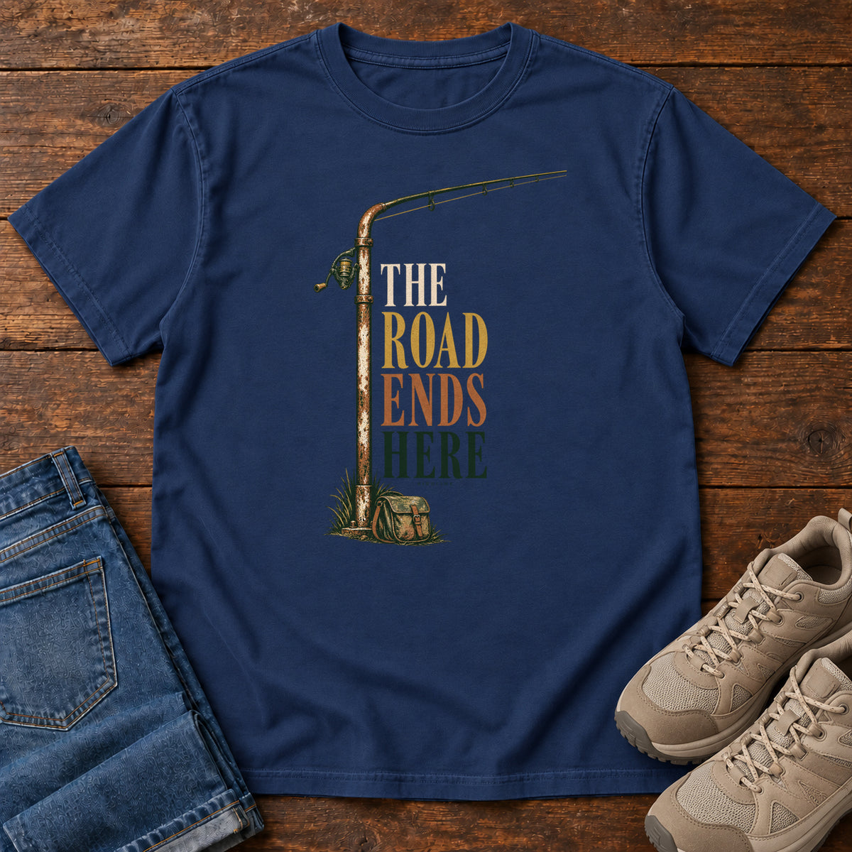 The Road Ends Here T-Shirt in True Navy, front laid flat on wood, featuring a fishing rod beside stacked earth-tone slogan lettering.