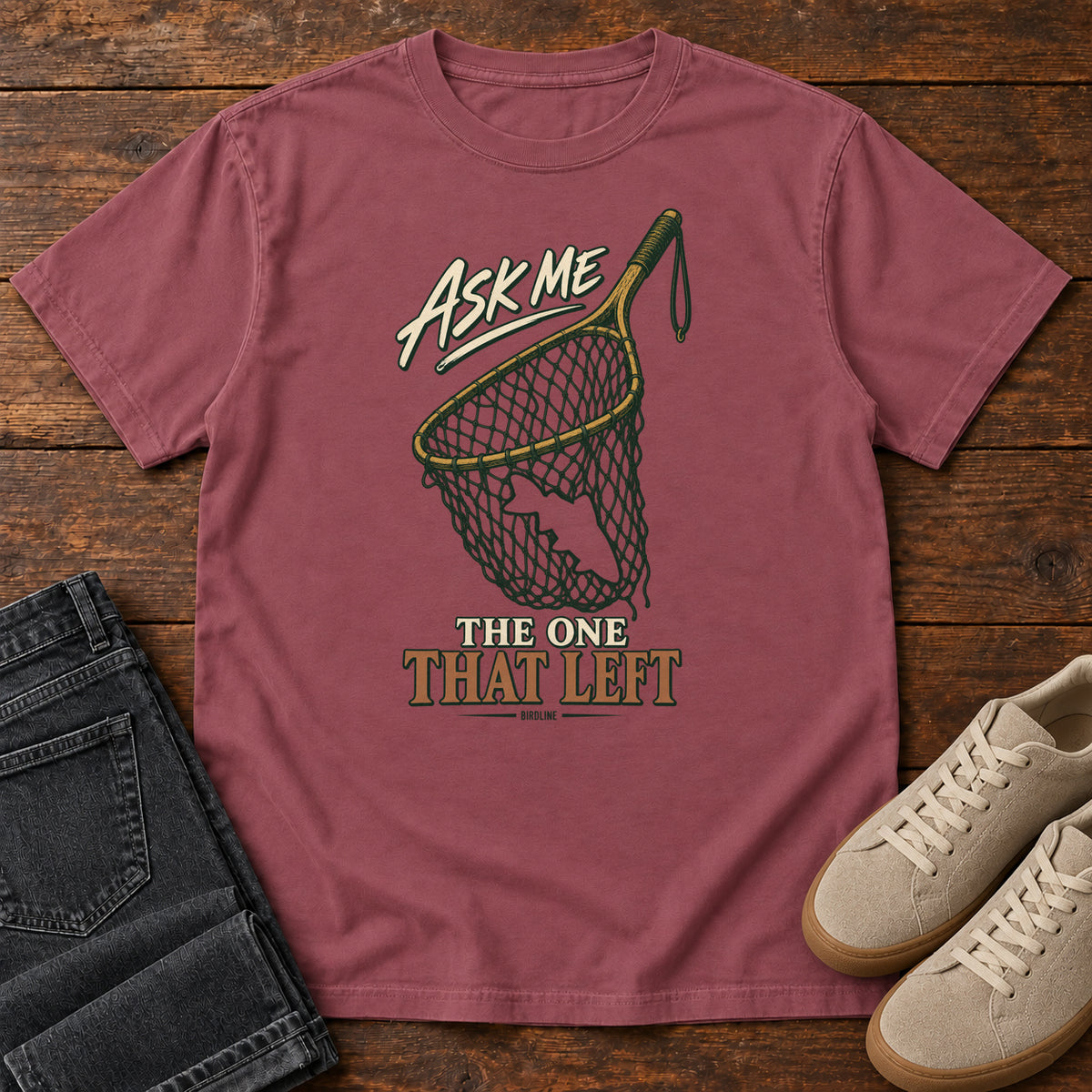 Brick Ask Me About the One That Left T-Shirt with an empty fishing landing net and cream-and-copper lettering, front flat lay on wood.