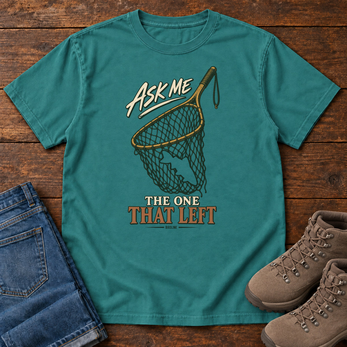Emerald Ask Me About the One That Left T-Shirt with an empty fishing landing net and cream-and-copper lettering, front flat lay on wood.