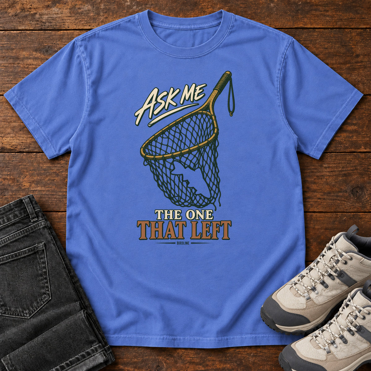 Mystic Blue Ask Me About the One That Left T-Shirt with an empty fishing landing net and cream-and-copper lettering, front flat lay on wood.