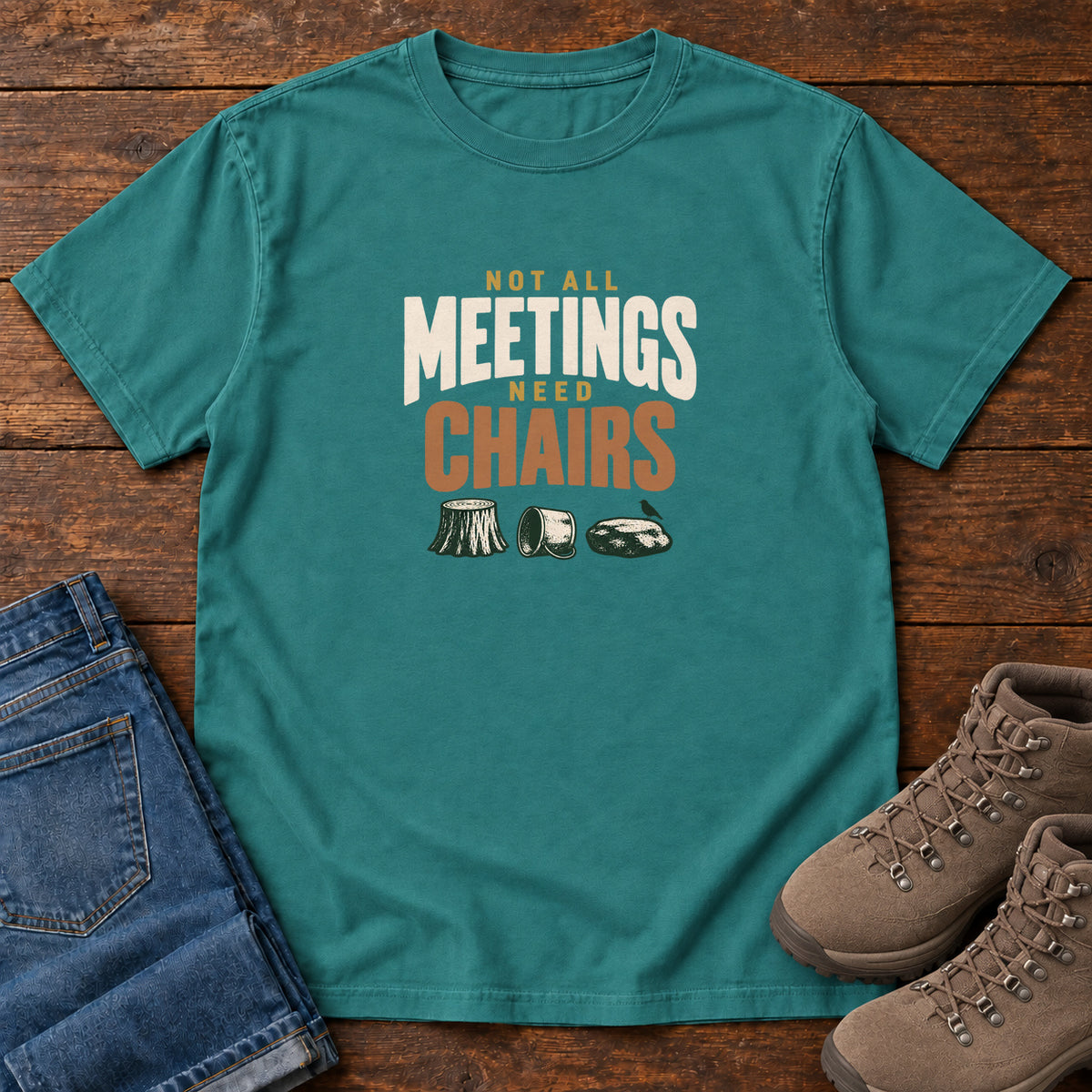 Emerald Not All Meetings Need Chairs T-Shirt with stacked cream-and-orange lettering above a stump, tipped mug and rock; front flat lay on wood.
