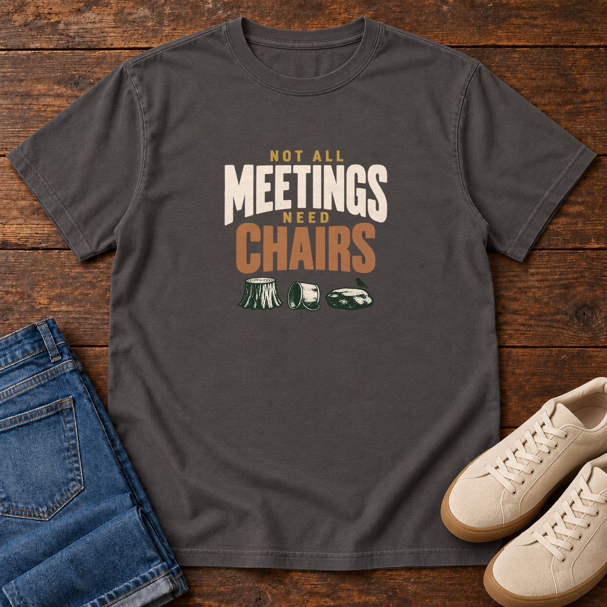 Graphite Not All Meetings Need Chairs T-Shirt with stacked cream-and-orange lettering above a stump, tipped mug and rock; front flat lay on wood.