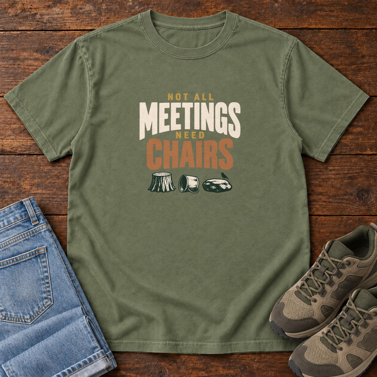 Moss Not All Meetings Need Chairs T-Shirt with stacked cream-and-orange lettering above a stump, tipped mug and rock; front flat lay on wood.