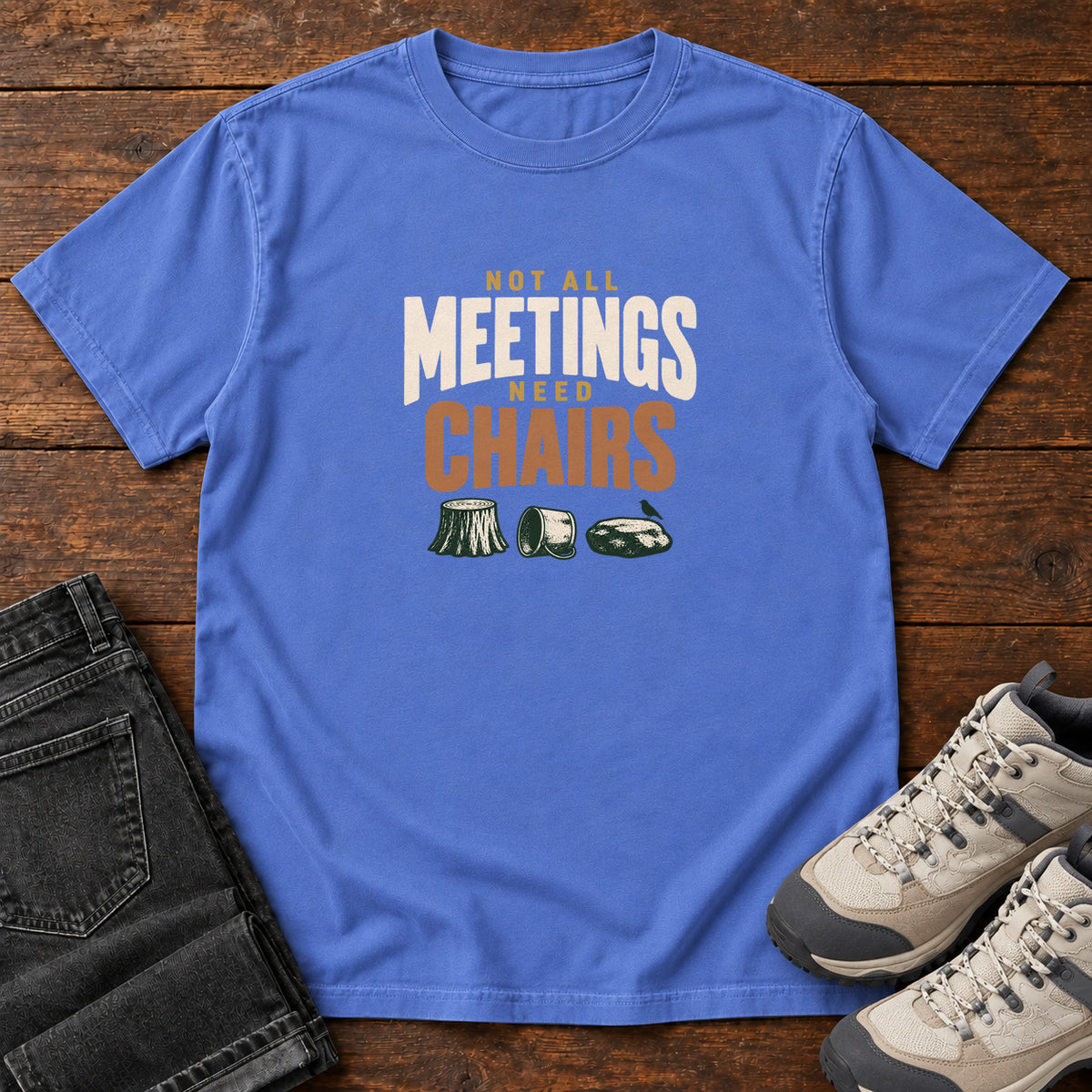 Mystic Blue Not All Meetings Need Chairs T-Shirt with stacked cream-and-orange lettering above a stump, tipped mug and rock; front flat lay on wood.
