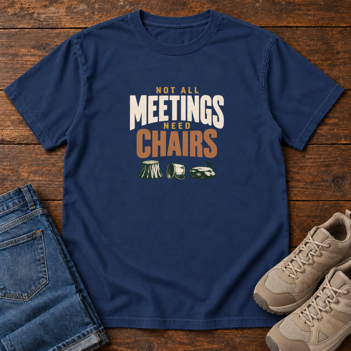 True Navy Not All Meetings Need Chairs T-Shirt with stacked cream-and-orange lettering above a stump, tipped mug and rock; front flat lay on wood.